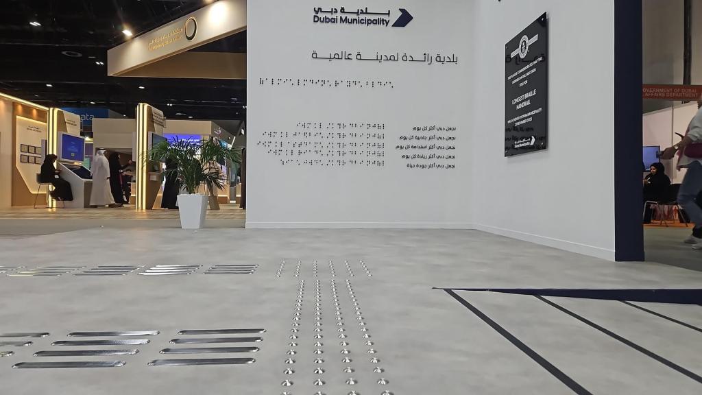 A view of a Dubai Municipality exhibition booth featuring a wall with Arabic and English text, surrounded by a modern exhibition space and attendees. The floor has metallic tactile strips for accessibility.