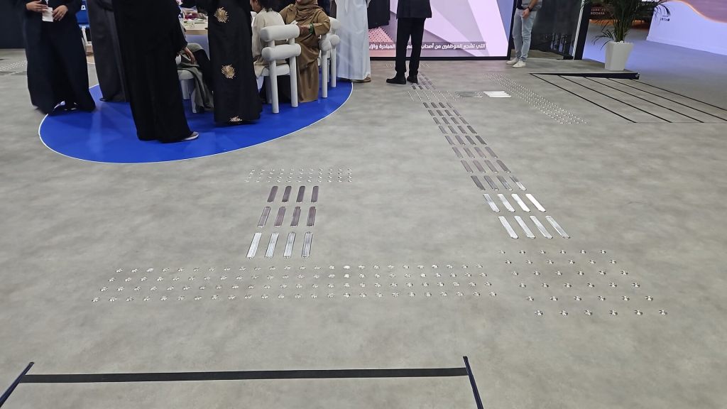 An exhibition space featuring decorative floor patterns made of metallic strips, with people standing around a round table in the background.