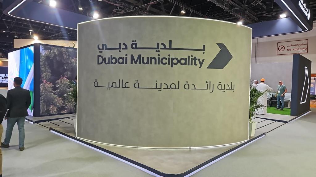 Exhibition booth for Dubai Municipality featuring the name in both Arabic and English, with decorative elements and attendees in the background.