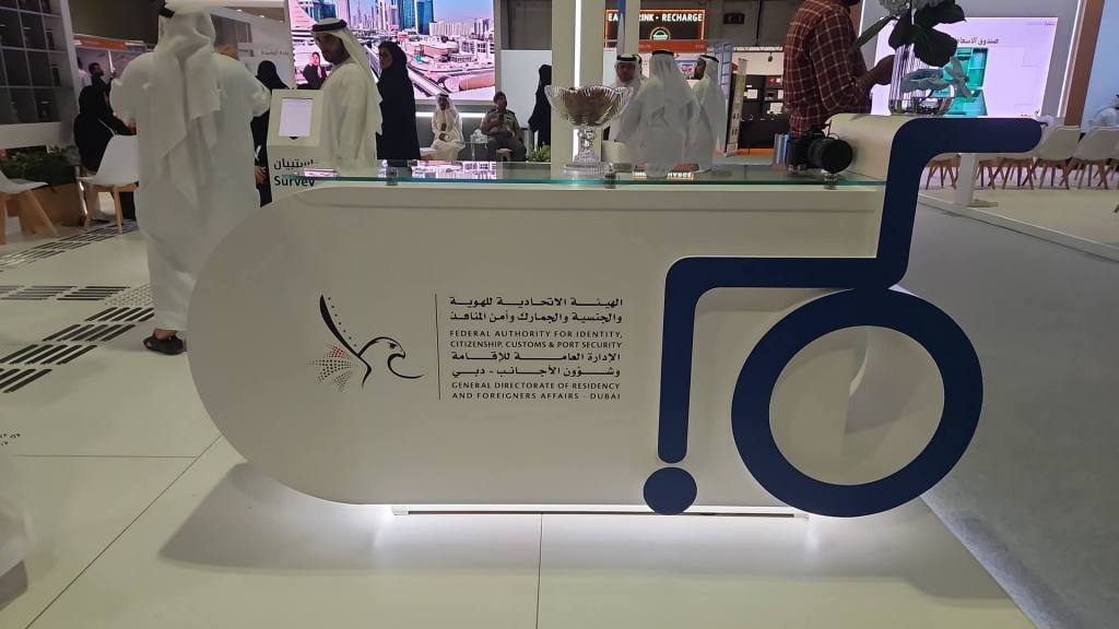 A modern reception desk featuring the logo of the Federal Authority for Identity, Citizenship, Customs, and Security, with a prominent accessibility symbol. The background shows people interacting at an event.