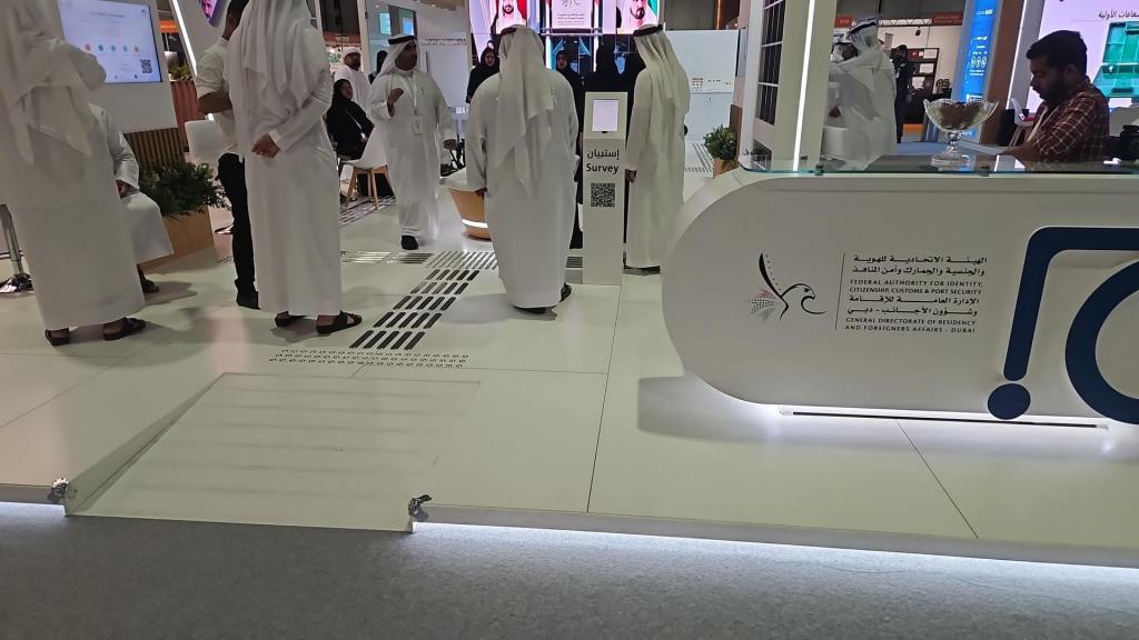 Exhibition space featuring a group of people in traditional attire, with a sleek modern booth displaying the Federal Authority for Identity and Citizenship. The area has a white floor and signage with a QR code for a survey.