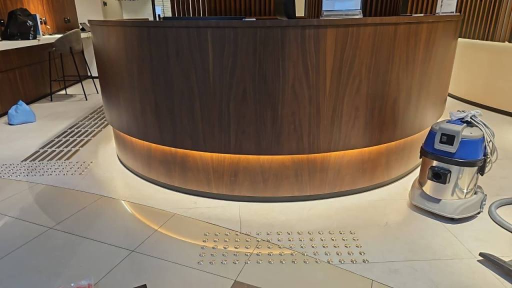 Image of a reception area featuring a wooden counter, stainless steel tactile studs installed on the floor, and a vacuum cleaner nearby.