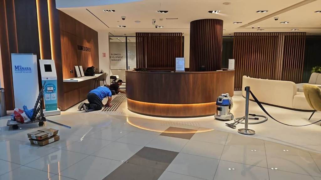 A worker installing stainless steel tactile studs on the floor of the Real Estate Registration Trustee LLC office in Al Manara Centre, Dubai. The reception area features modern design elements and a sleek counter.