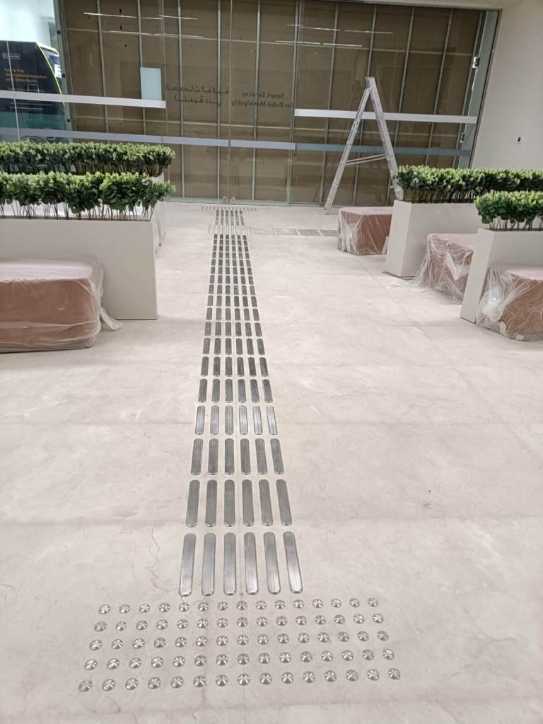 Installation of tactile stainless steel strips and studs on a polished floor, with planters and seating areas visible in the background.