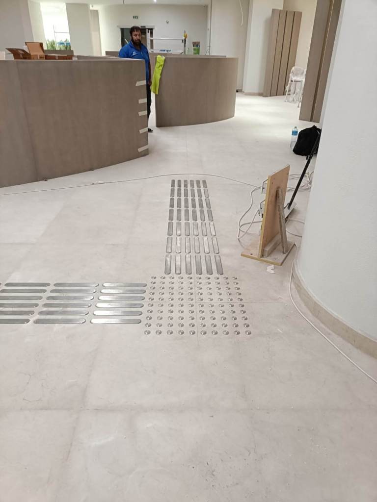 Installation of tactile stainless steel strips and studs on floor at Dubai Court, with worker visible in background.
