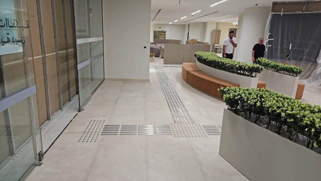 Interior of the Dubai Court at Manara Mall, featuring tactile stainless steel self-adhesive strips and studs installed in a corridor, with decorative planters and furniture in the background.