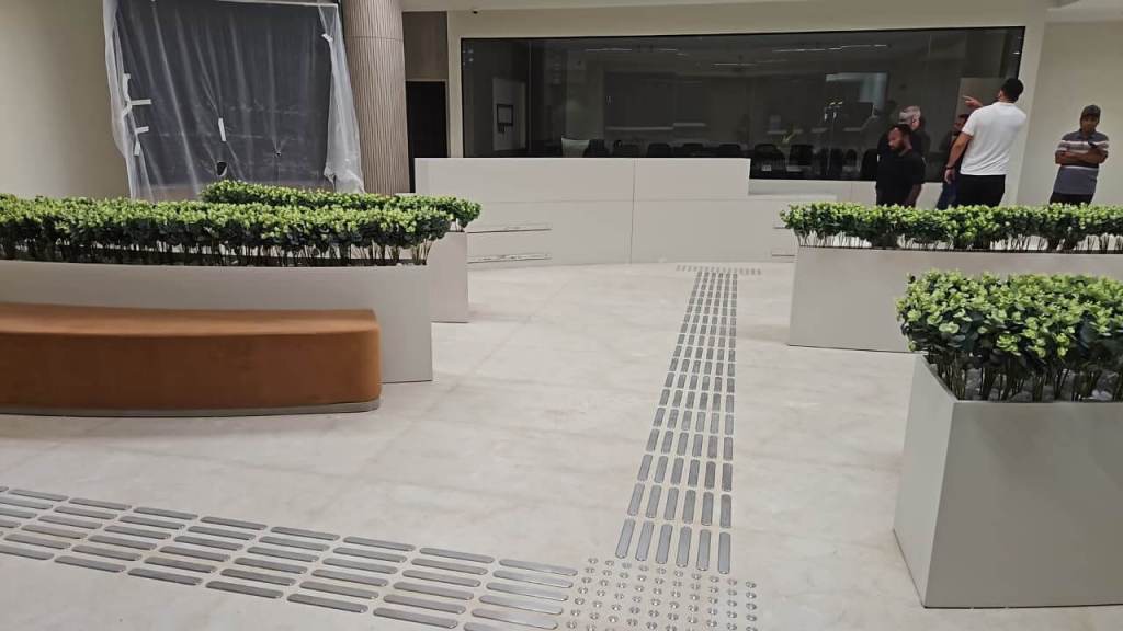 Interior view of Dubai Court at Manara Mall featuring tactile stainless steel strips and studs integrated into the flooring, surrounded by potted greenery and seating areas.