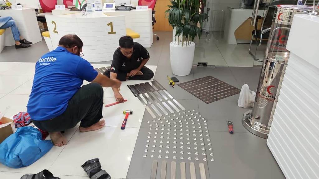 Two workers install stainless steel tactile studs on the floor in an office space, aligning materials for accessibility.