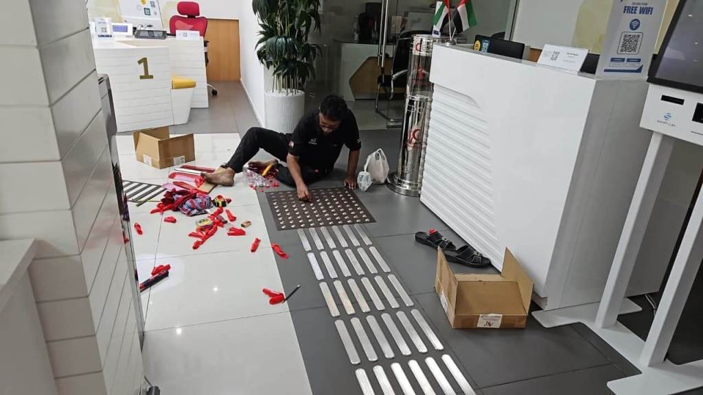 A technician installing stainless steel tactile self-adhesive studs and lines on the floor of an office in Dubai. Tools and materials are spread out around him on the polished tile floor.