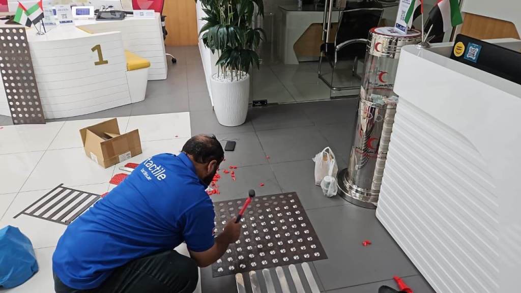 A worker in a blue shirt installs stainless steel tactile studs on a tiled floor in an office lobby, with an assortment of tools and materials scattered around.