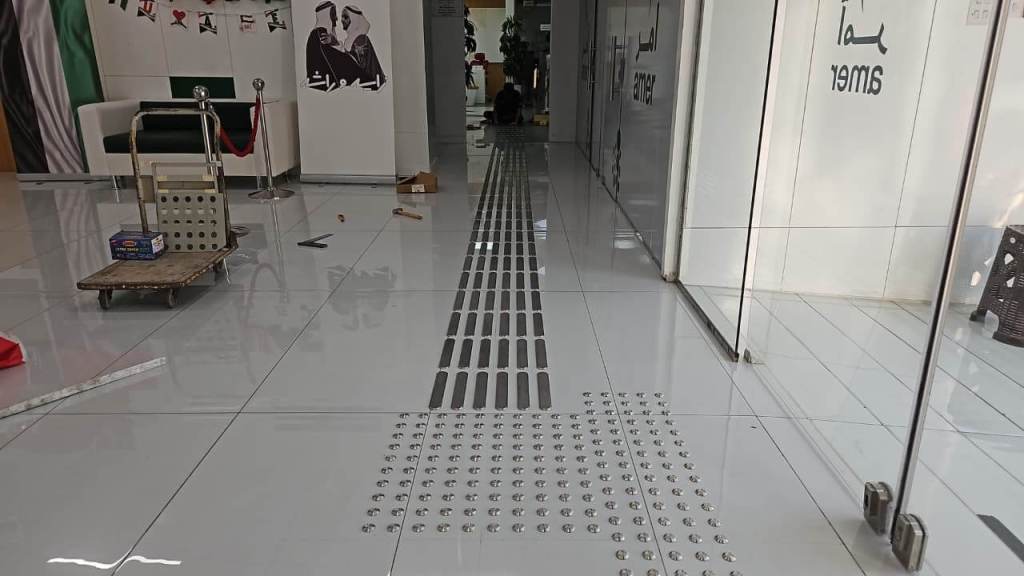 Installation of stainless steel tactile self-adhesive studs and lines on polished tile flooring in an office environment, showcasing accessibility features.