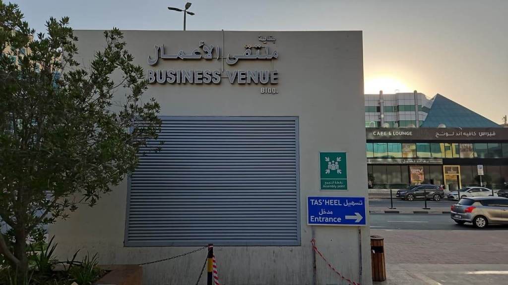 Signage for Business Venue in Dubai, with text in Arabic and English. Features surrounding greenery and buildings in the background, hinting at a busy commercial area.