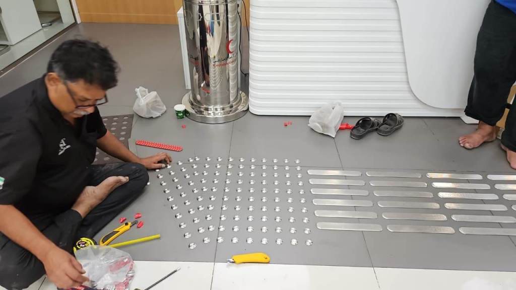 A worker installing stainless steel tactile studs on a floor, with tools and components laid out nearby.