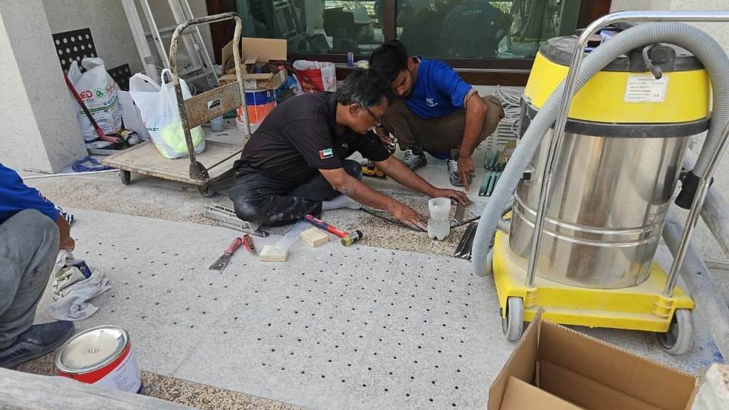 Two workers installing stainless steel tactile pin-type studs on a concrete surface, surrounded by tools and materials.