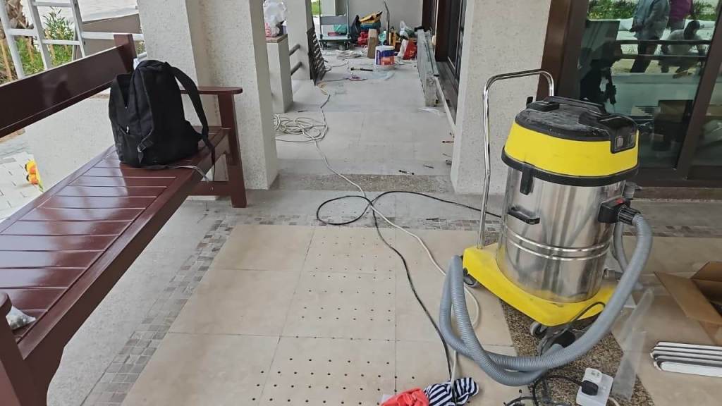 Image showing a construction site at Majlis Al Bahar, Jumeirah. A brown bench is positioned on the left, alongside a black backpack. In the foreground, there's a gray and yellow vacuum cleaner. The area is under renovation, with construction materials and tools visible in the background.