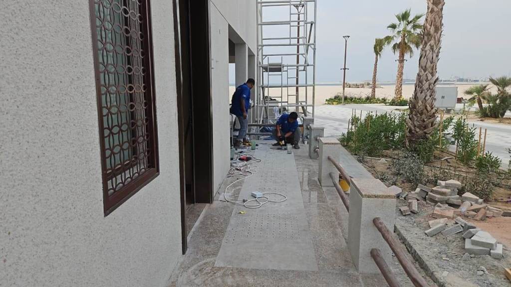 Two workers installing tactile paving studs at the entrance of the Majlis Al Bahar building in Jumeirah, with construction materials and equipment visible in the foreground.