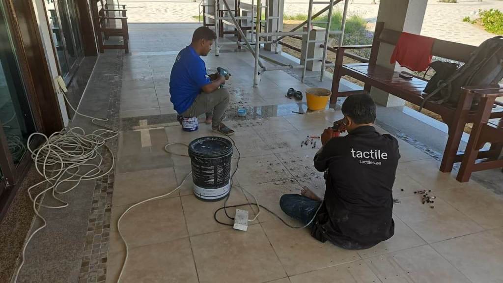 Two workers installing tactile indicators on tiles in a semi-outdoor area. One is operating a tool while the other is preparing materials, with tools and equipment visible around them.