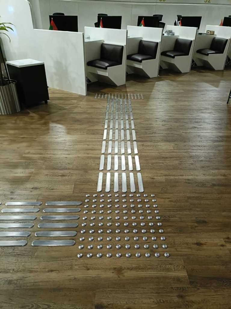 A view of a modern workspace with white partitions and dark seating. The floor features a pattern of metal strips and round markers arranged in organized rows.