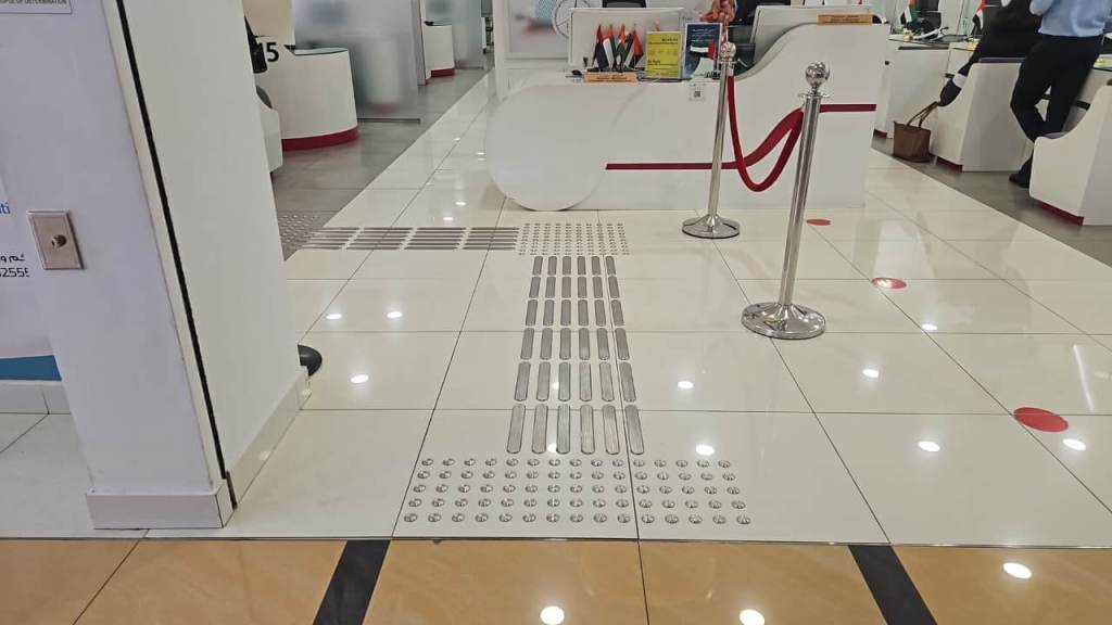 A tiled floor with tactile guidance strips and raised dots for accessibility, leading to a service area with counters and stanchions in a bright interior space.
