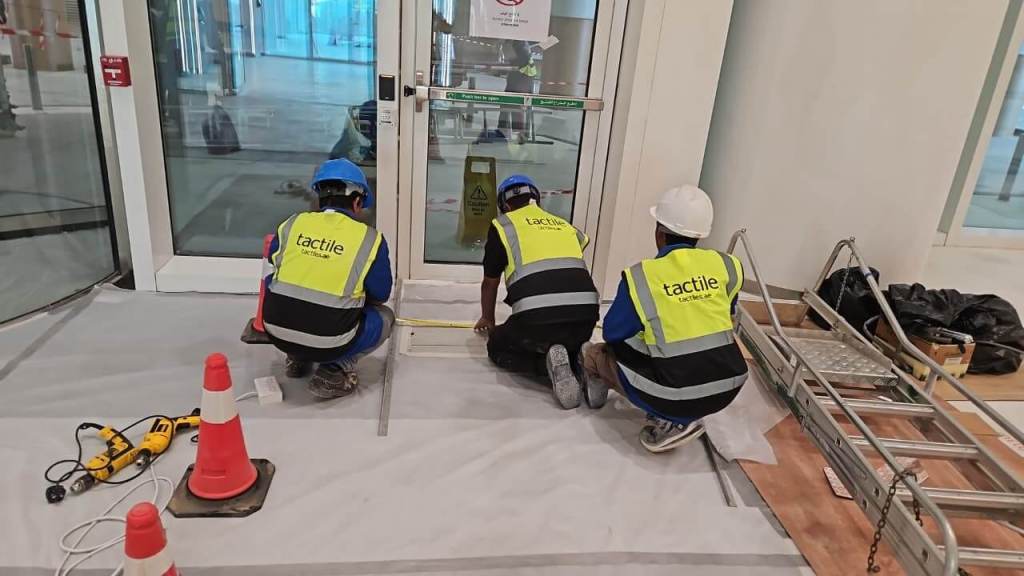 Three construction workers in safety vests are working on the floor near a glass door, measuring and preparing the area.