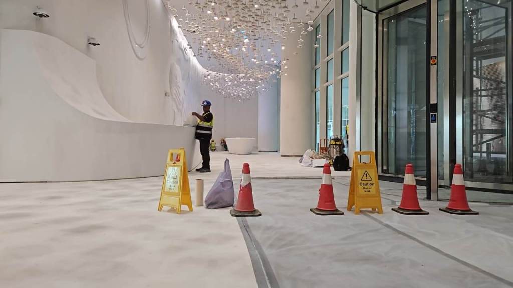 A construction site indoors featuring a worker in a striped uniform, caution signs, and traffic cones on a white surface. Decorative lighting hangs from the ceiling.