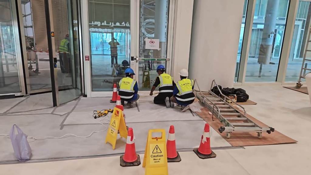 Three construction workers in safety vests are performing tasks near a glass entrance, surrounded by caution cones and warning signs. A ladder and tools are visible in the area.