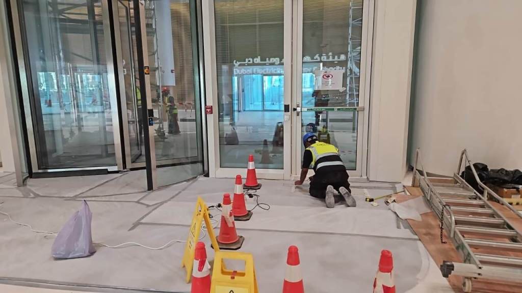 A construction worker in a safety vest kneels on the floor near double glass doors, which are flanked by construction cones and caution signs. The area appears to be under renovation, with tools and materials visible in the background.