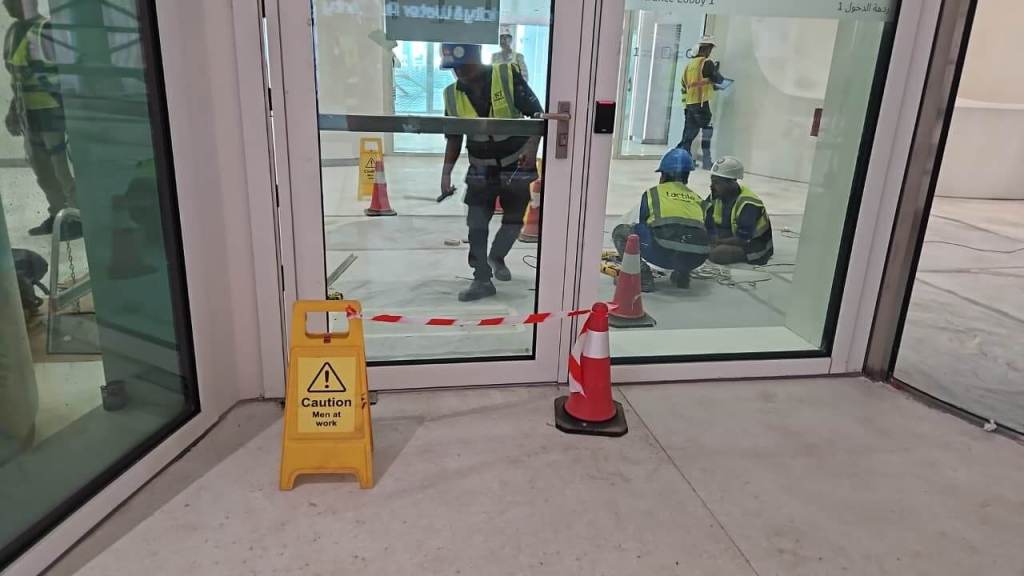 Construction workers wearing safety vests and helmets inside a building under renovation, with caution signs and cones present near a glass door.