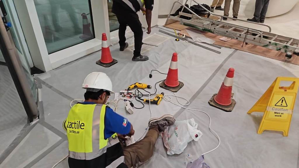 A construction site with a worker in a safety vest and helmet sitting on the floor, using tools and electrical cords. Caution cones and a wet floor sign are visible in the area.