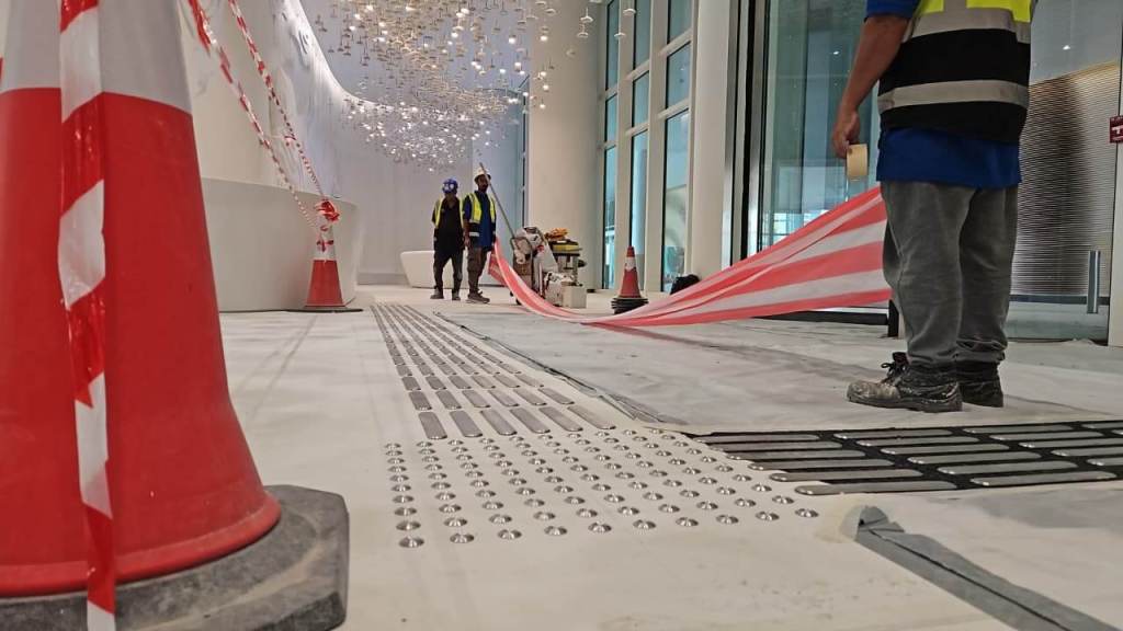 Construction workers setting up safety tape in a newly renovated indoor space, with barriers in place and decorative ceiling lights visible.