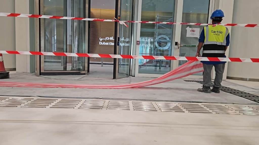 A worker in a blue helmet and safety vest stands near a construction area with red and white caution tape blocking the entrance to a building. The floor is under construction, and there is a sign visible on the door.