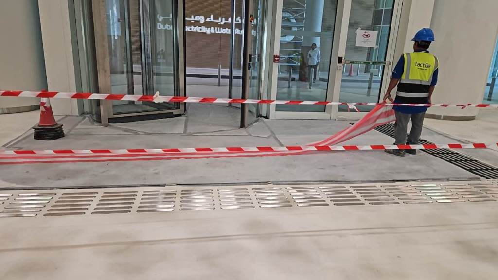 A construction worker in a blue helmet and reflective vest stands near a barrier of red and white caution tape. The entrance to a glass door is visible in the background, with a sign indicating ongoing work.