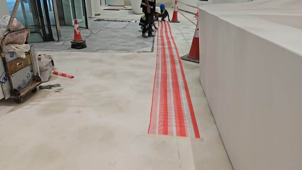 Construction workers laying down red and white hazard tape on a floor in a commercial space under renovation, with traffic cones and construction equipment visible.