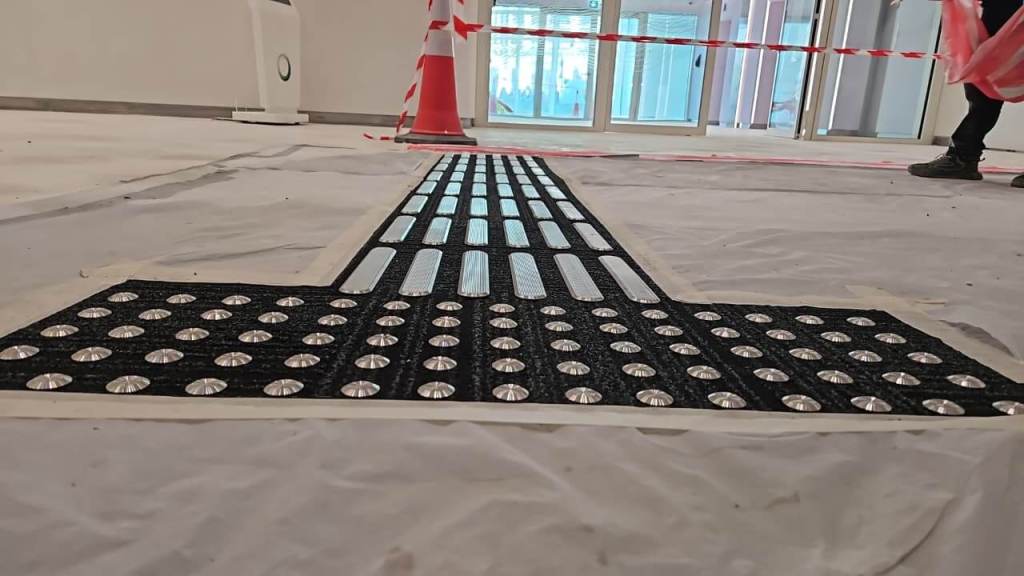 Close-up of a tactile ground surface indicator with studs, placed on a construction site floor covered with plastic sheeting. Traffic cones and caution tape are visible in the background.