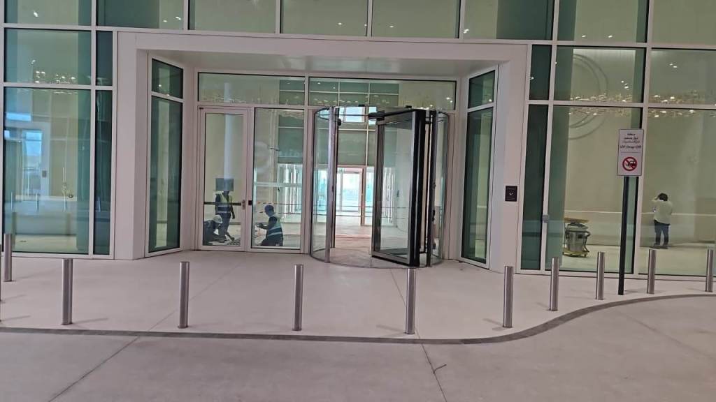 Entrance to a modern building featuring a glass revolving door, large windows, and a smooth concrete pathway with bollards.