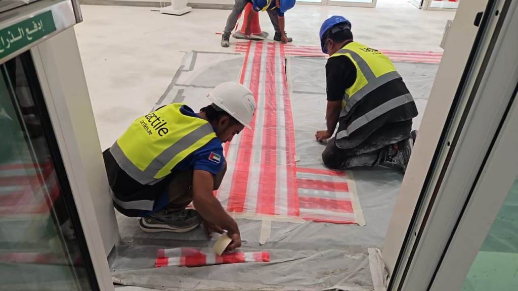 Two construction workers wearing safety vests and helmets are laying down red and white tape on a floor covered with protective sheeting in a partially completed interior space.