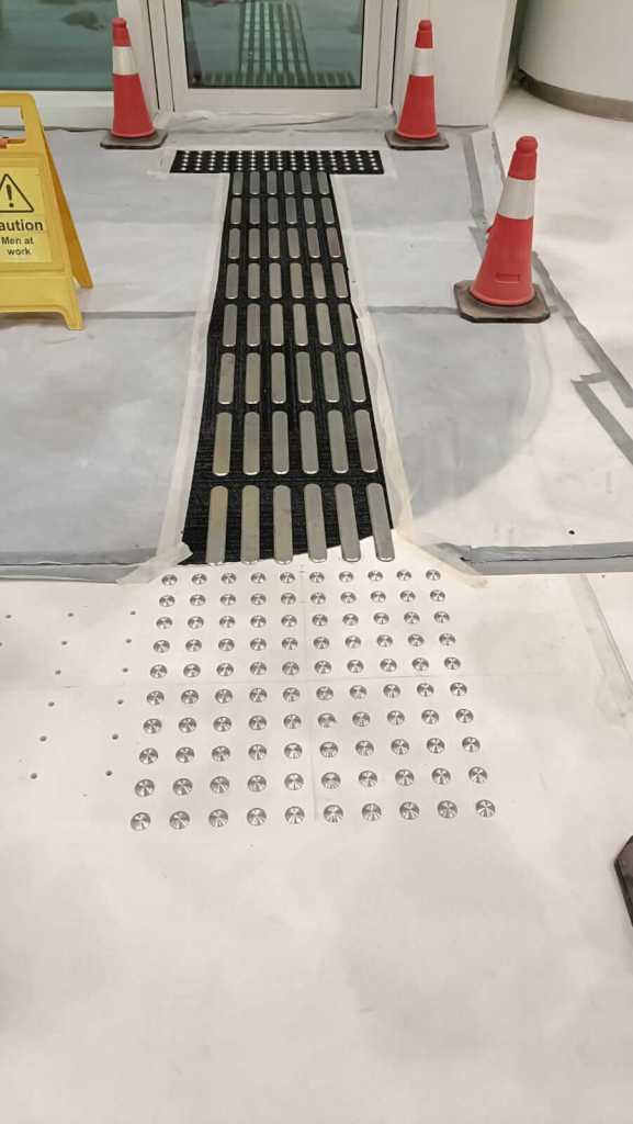 A pathway with textured strips and tactile paving leading to a door, surrounded by orange traffic cones and a caution sign indicating construction work.