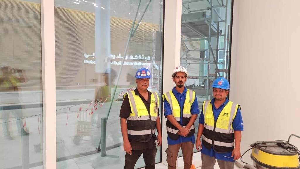 Three construction workers wearing safety vests and helmets pose for a photo inside a building under construction, with scaffolding and tools visible in the background.