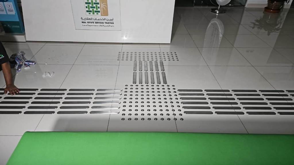 A close-up of a tiled floor featuring a pattern of raised and recessed elements, with a person's hand reaching towards it. A green bench is partially visible in the foreground.