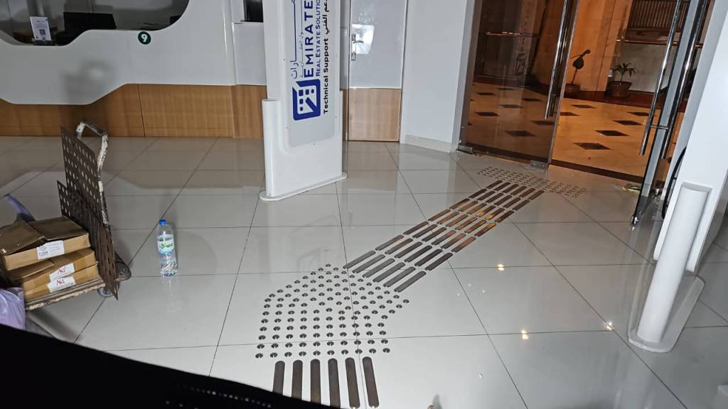 Interior view of a lobby featuring a patterned floor with tactile guidance strips, a standing sign for Emirates Technical Support, and various items on the floor including boxes and a water bottle.