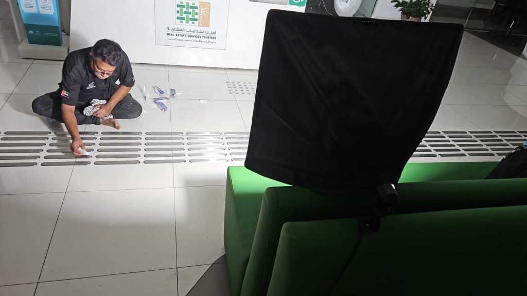 A person wearing a black shirt is working on the floor, focusing on a row of ventilation ducts. Tools and materials are scattered around, and a green couch is visible in the foreground.