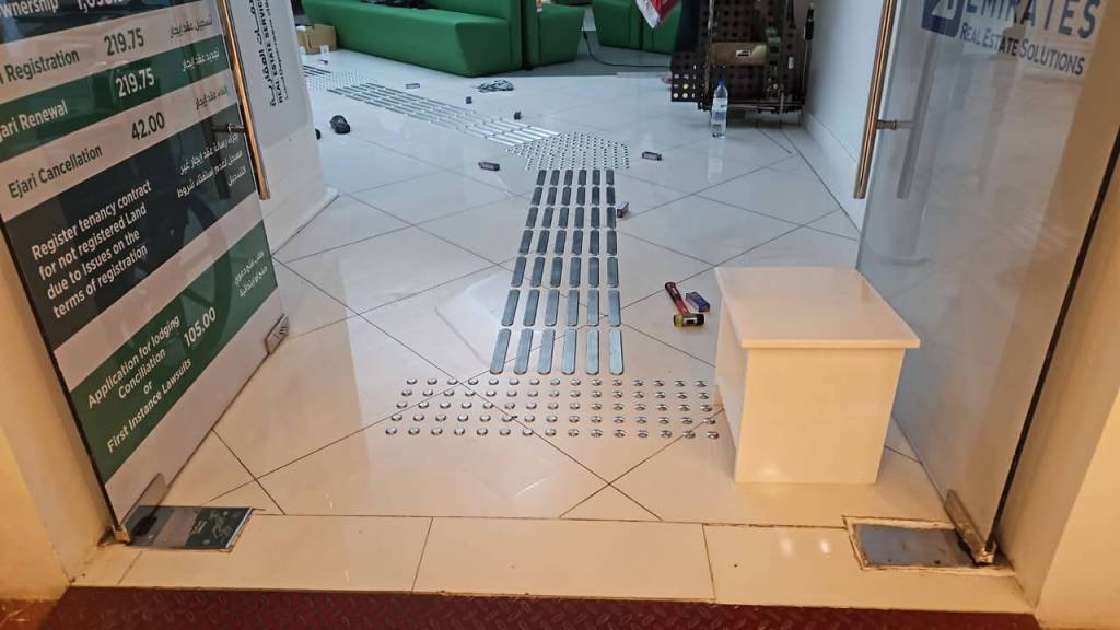 Interior view of a reception area featuring tiled flooring, a green seating area in the background, and a white storage box near the entrance. The floor has metal strips and markers indicating a pathway.