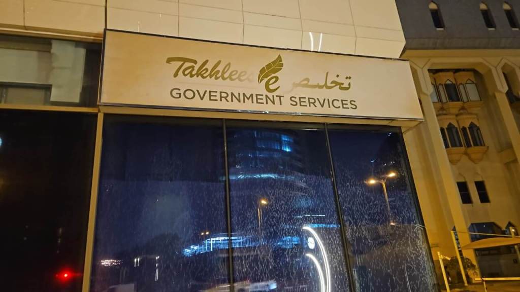 Sign for 'Takhlilee Government Services' in Arabic and English, illuminated at night with rain on the glass facade.
