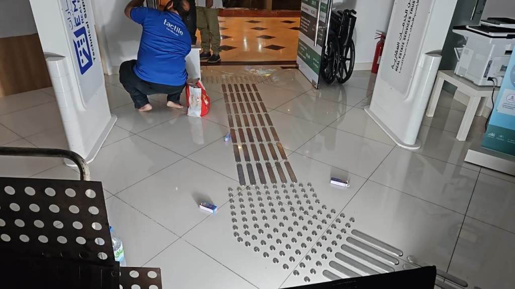 A person in a blue shirt is kneeling on the floor near a tactile guiding path in a lobby-like space. The floor is tiled with visible signage and a wheelchair in the background.
