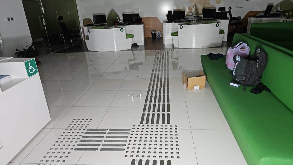An office interior featuring a green couch, reception desks, and a tactile paving strip on the floor. There is a box and a backpack on the floor.