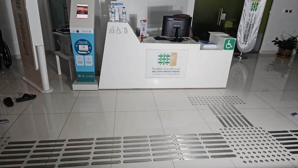 Reception area with a modern counter displaying a logo and accessibility signs. A desktop computer and water bottles are visible on the counter, along with decorative plants in the background.