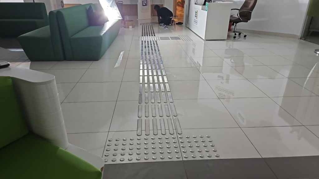 A brightly lit indoor space featuring glossy white tiles and green seating. Visible is a tactile guidance pathway with textured strips and raised dots for accessibility. A person is working on the floor near the reception area.