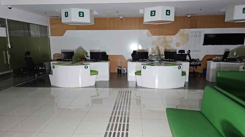 Interior view of a modern office reception area with white desks and green chairs. The space features numbered service counters and a glass wall on one side. Brightly lit with a clean, minimalist design.