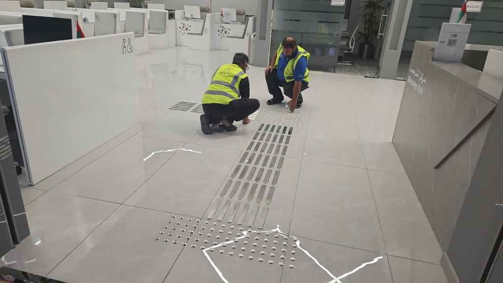 Two workers in safety vests inspecting floor drainage grates in an office space.