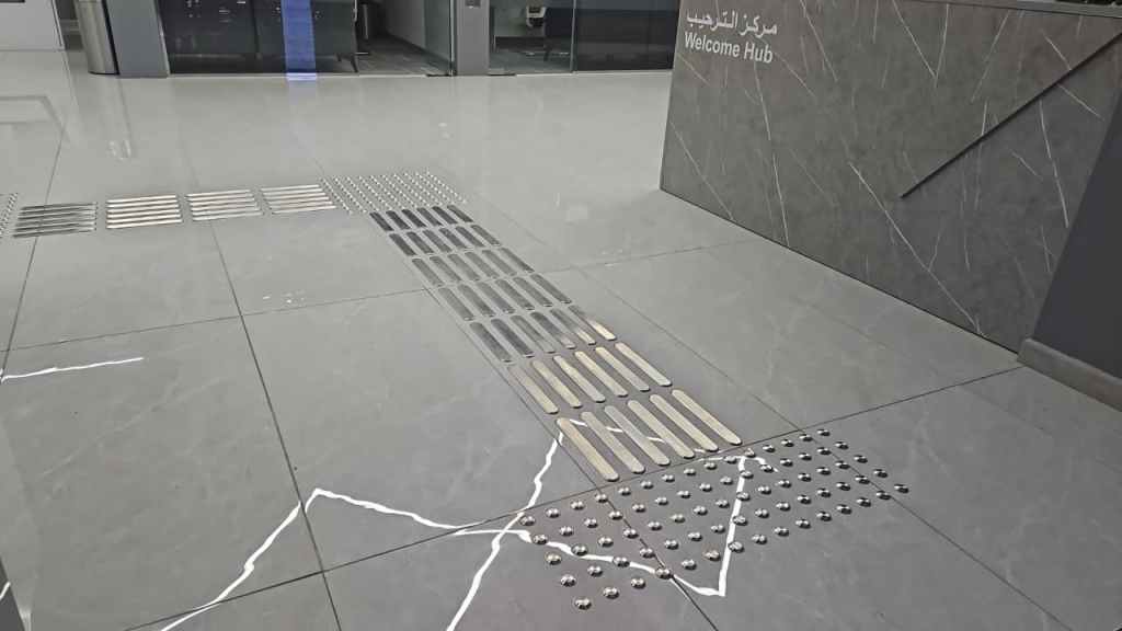 Interior view of a modern building showcasing a sleek gray floor featuring textured pathways, including strips and dots, leading towards a 'Welcome Hub' sign in both Arabic and English.